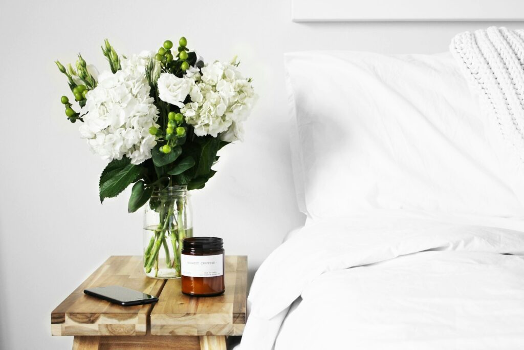 flowers in clear glass vase on table next to a clean white bed