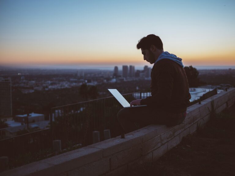 digital nomad sitting on ledge overlooking his new city with opened laptop on his lap